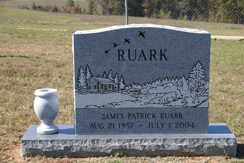 Grey upright monument etched with a cabin, pines, deer and geese in flight